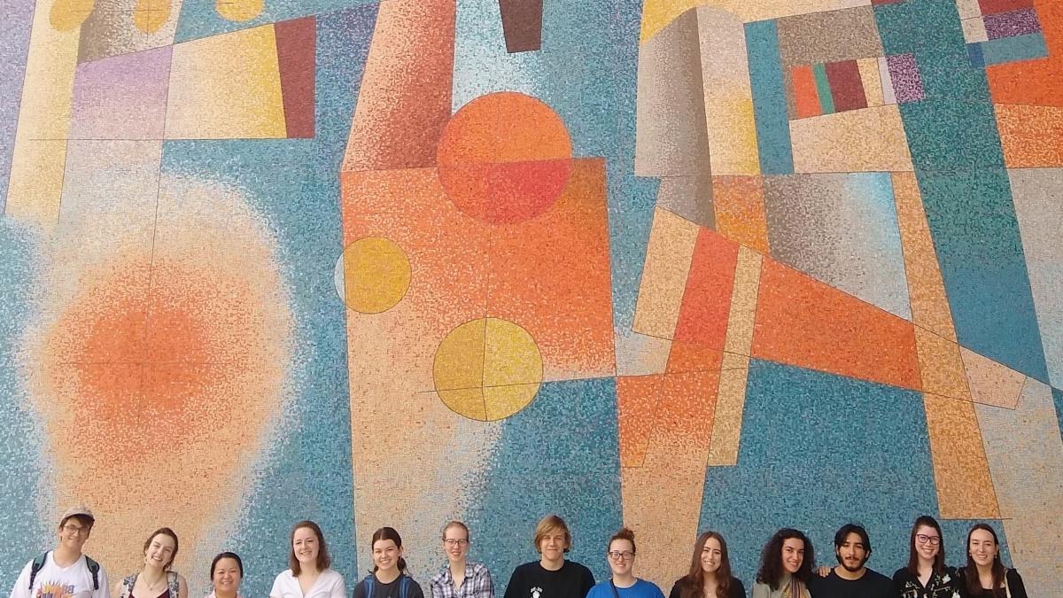 students stand collectively in front of a large glass mosaic by Carlos Merida